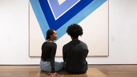 Two people in an art gallery sitting in front of a painting