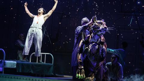 A man stands on a bed onstage next to a tiger puppet in the A.R.T.'s production of Life of Pi